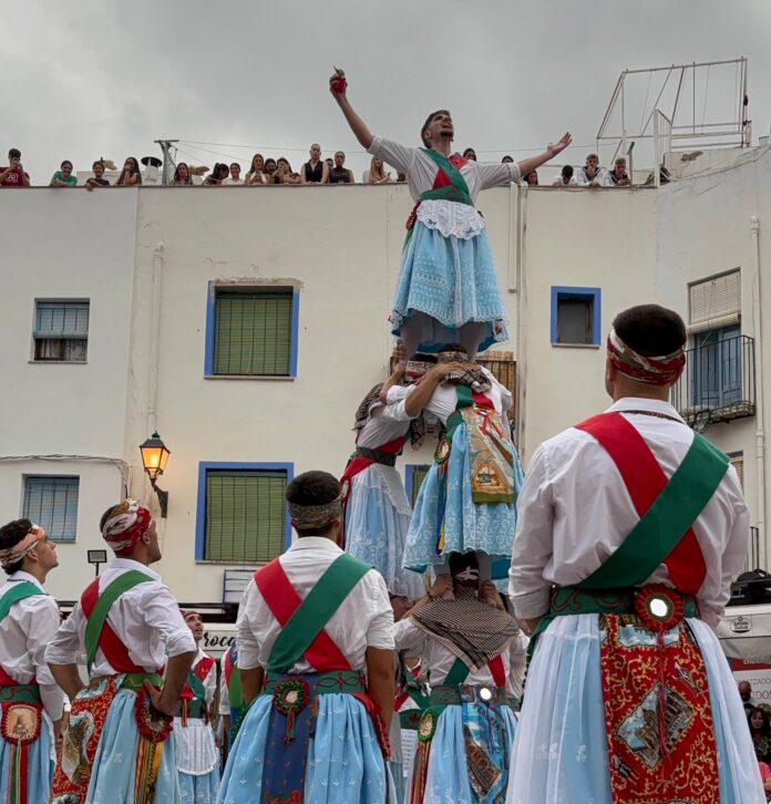 Group of performers in matching blue dresses with red/green sashes carry a man on shoulders during a street festival.