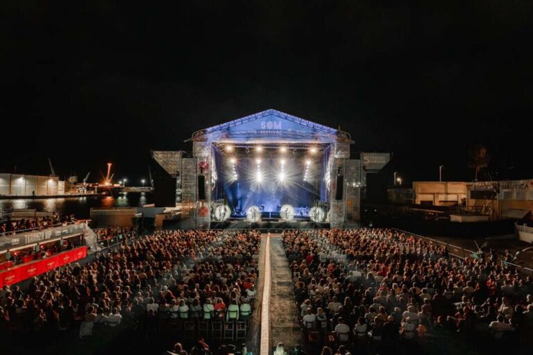Outdoor night concert with a lit stage and a large audience seated in rows by the water's edge at a festival venue