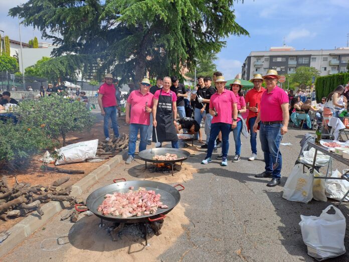 Group of people in pink shirts and straw hats grilling meat on large pans at an outdoor street barbecue.