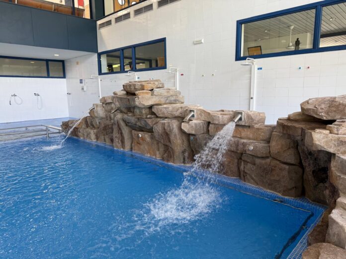 Indoor pool with a stacked rock waterfall feature and water jets spilling into a blue-tiled pool, white tiled walls, and shower stations nearby