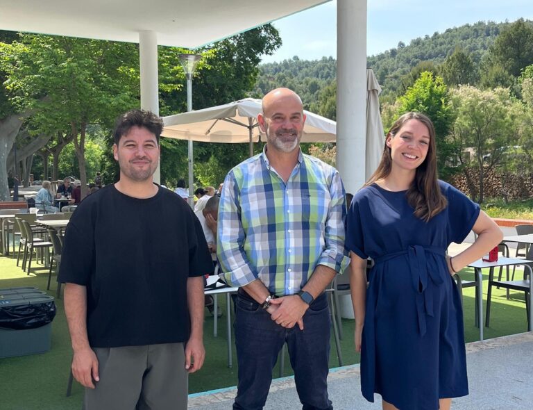 Three people stand together outdoors under a white canopy, smiling, with tables and green hills in the background.