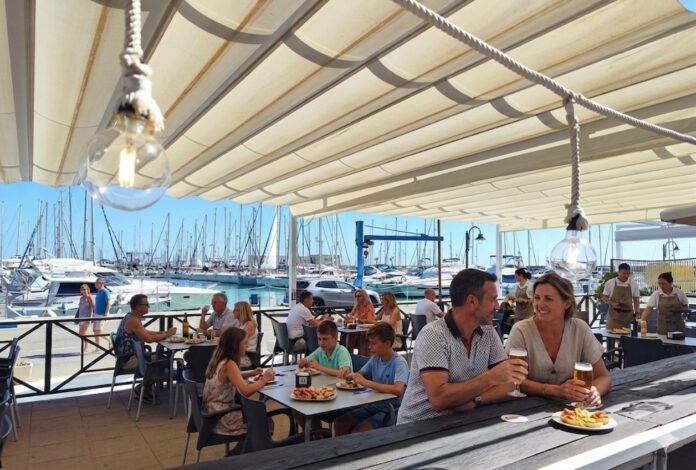 Couple with beers at an outdoor marina restaurant, plates of food in the foreground, sailboats in the harbor behind.