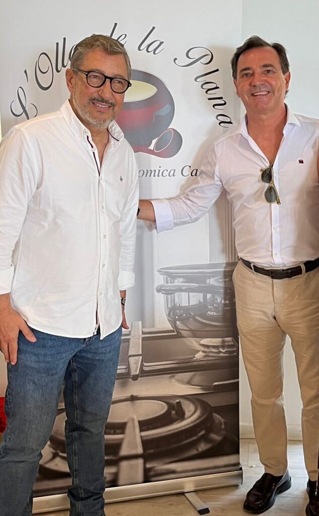 Two men in white shirts smile and pose together in front of a wine-themed banner with barrel imagery.