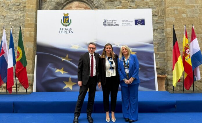 Three adults in business attire pose for a photo on a blue stage with a European flag backdrop and country flags on either side, at an EU/CERAMICS event.