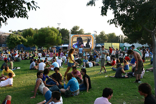 Crowd sitting on the grass at a sunny outdoor festival in a park; a large inflatable screen with performers visible in the background.