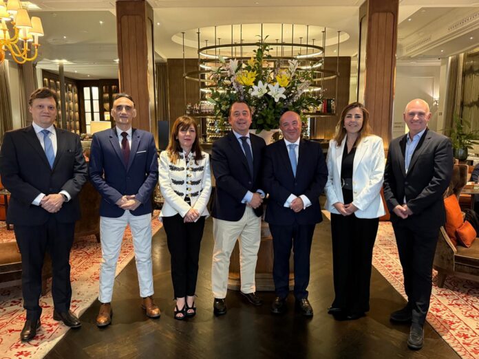 Seven professionals in business attire posing in a hotel lobby with a large floral arrangement behind them.