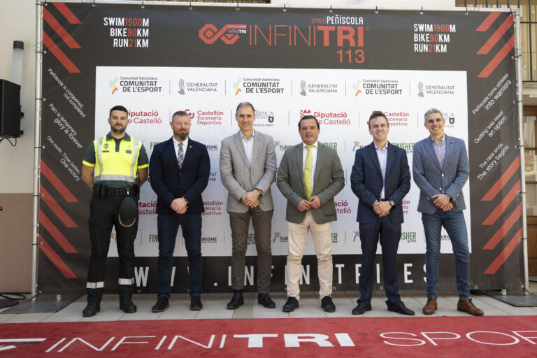 Five men in suits and one in a high-visibility vest stand on a stage in front of a branded backdrop at an event.