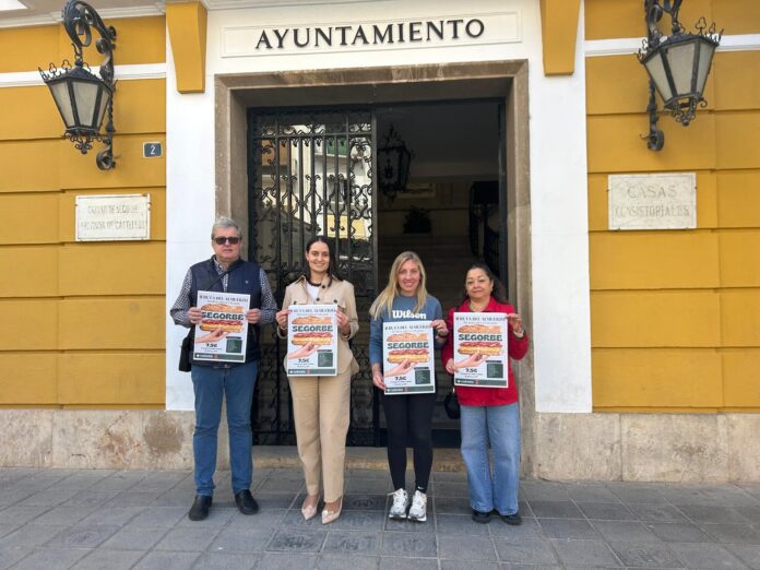 Four people stand in a row outside a yellow city hall, each holding a poster advertising Segorbe.