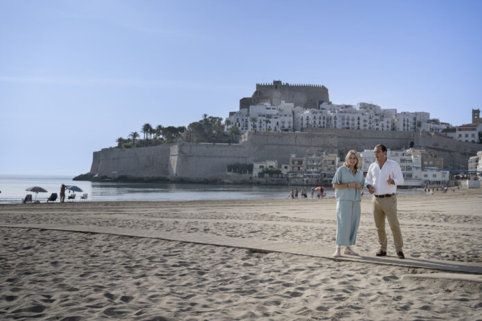 Couple walking on a sandy beach with a historic stone fortress and white hillside buildings in the background under a blue sky.