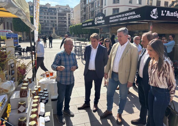 Vendor explains jars of honey on a display table to a group of adults in a city plaza street market.