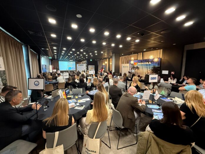 A large conference room with round tables filled with attendees listening to a presentation at the front stage, screens and banners visible in the background.