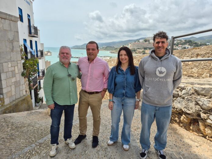 Four adults standing together on a stone walkway overlooking a sea town, smiling at the camera with blue-roofed buildings in the background.