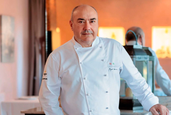 Professional chef in a white jacket standing in a warm, well-lit kitchen with stainless steel equipment in the background.