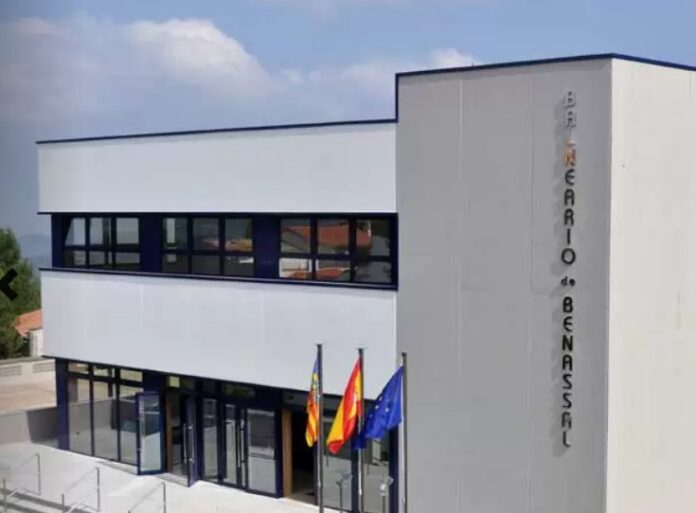 Modern two‑story building with a glass entrance and a vertical sign on the right, three flags (regional, national, and EU) fluttering in front under a blue sky.