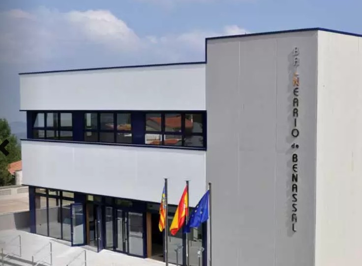 Modern two‑story building with a glass entrance and a vertical sign on the right, three flags (regional, national, and EU) fluttering in front under a blue sky.
