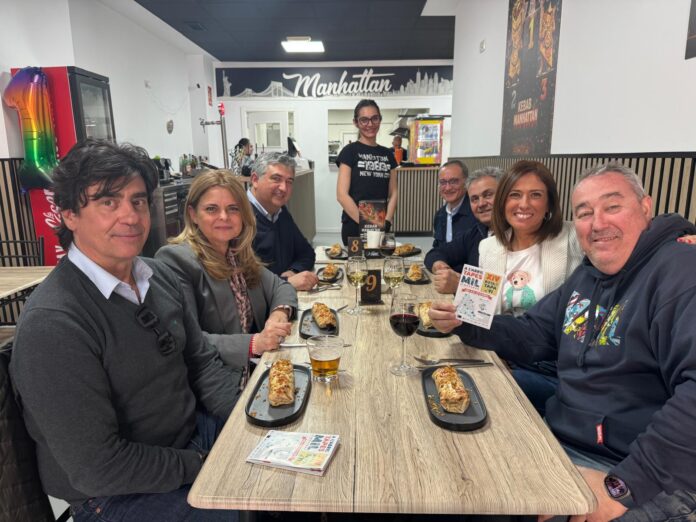 Group of eight adults seated at a restaurant table, smiling, with pastries and drinks; a waitress stands in back near a 'Manhattan' sign.