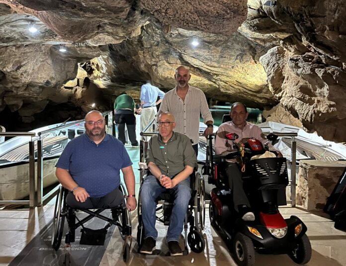 Group of people in wheelchairs along a glass walkway inside a dimly lit rocky cave, with a red mobility scooter nearby and rock walls above.