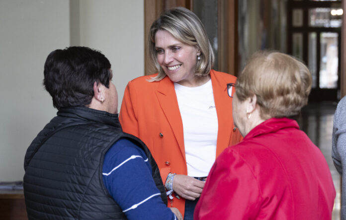 Smiling woman in an orange blazer talks with two people in a hallway or lobby setting.