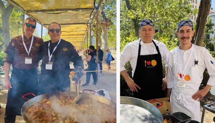 Two men in sunglasses and lanyards stand beside a large steaming outdoor pot at a sunny cooking event.