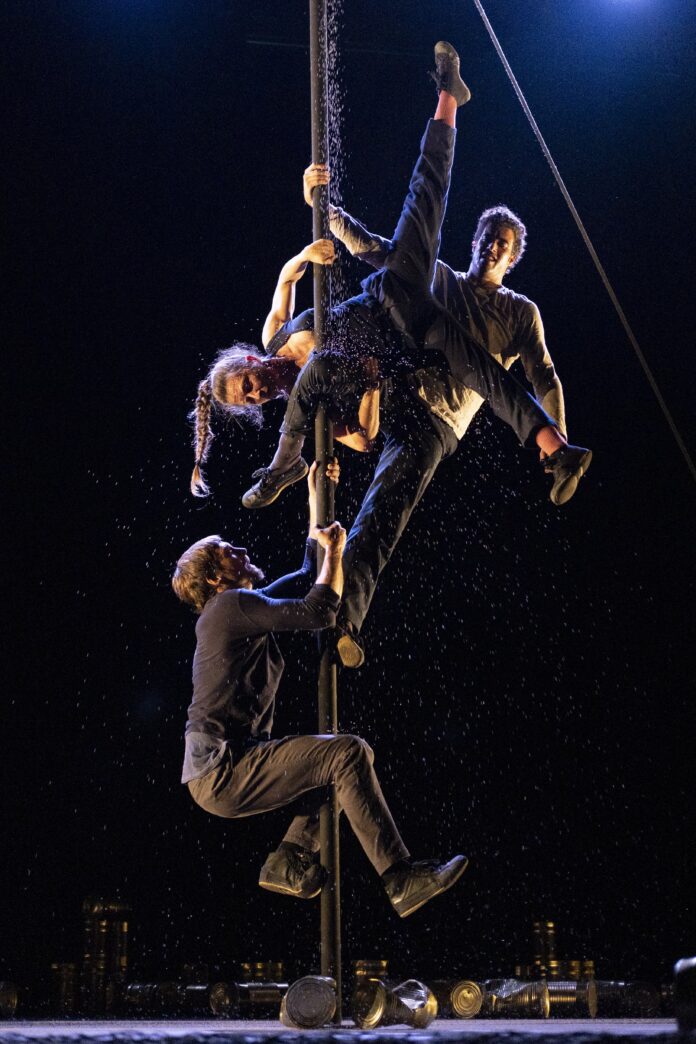 Climbers perform on a tall pole at night, suspended by hands and feet with water droplets falling around them at ground level lights in the distance.