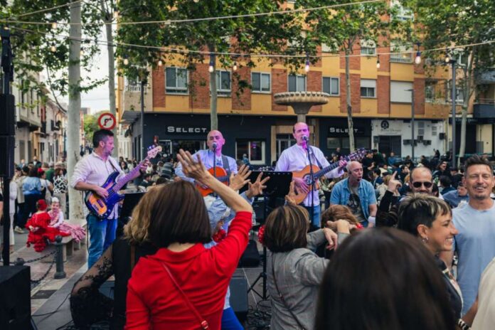 Band performs live on an outdoor street stage as a crowd dances and raises hands.