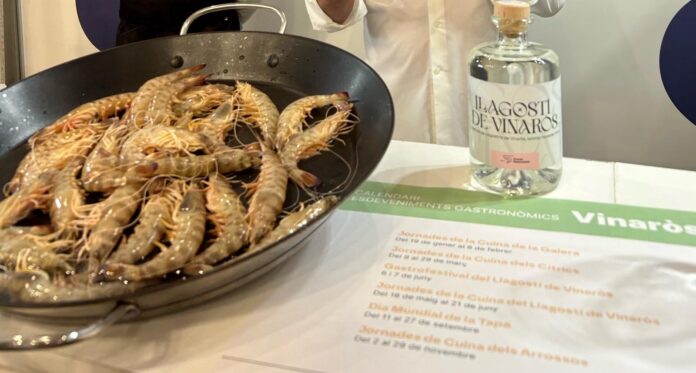 Pan filled with raw shrimp and prawns on a black skillet, next to a bottle labeled Il Lagost de Vinaròs and a festival brochure on a table.