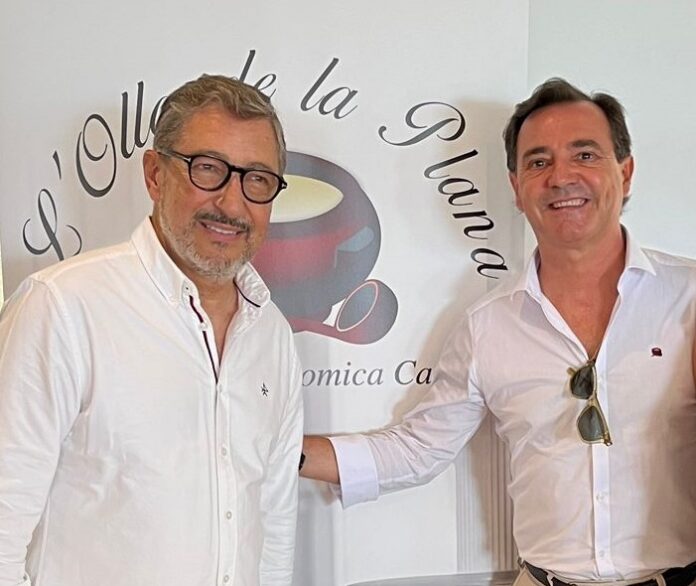 Two men in white shirts smile at the camera in front of a logo banner for a café or brand event.