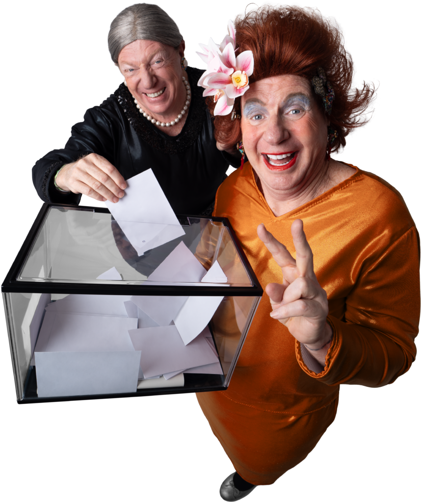 Two older women in festive costumes vote, placing papers into a clear ballot box.