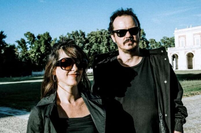 Two adults in black jackets and sunglasses stand outdoors on a sunny day, with trees and a light-colored building in the background.