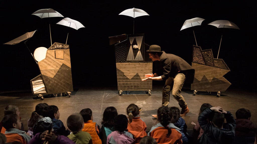 Performer in a hat leans toward seated children on a dark stage, surrounded by abstract wooden sculptures and white umbrellas on wheels.