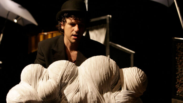 A man in a black hat and blazer on a dim stage with large white rope-like sculptures in the foreground.