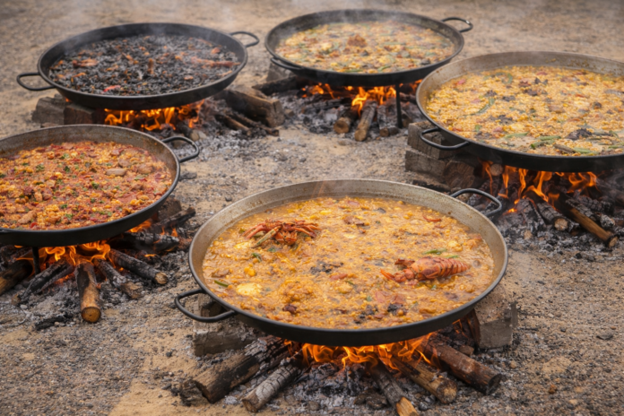 Five large frying pans simmer over an outdoor campfire, cooking different stews and paellas with visible vegetables and meats.