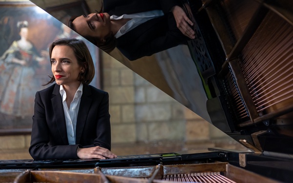 Professional woman in a dark blazer sits at a grand piano, hands folded, in a recital hall; her reflection is visible on the piano lid.