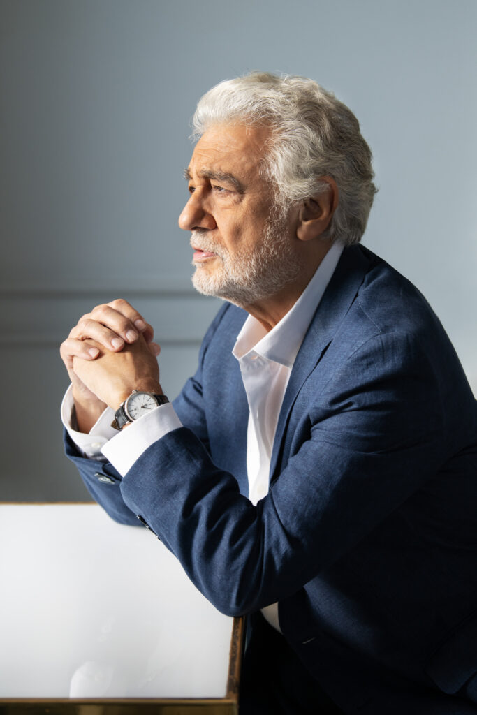Older man with white hair and beard in a blue suit, seated at a desk with hands clasped in front of him, looking thoughtful to the left.