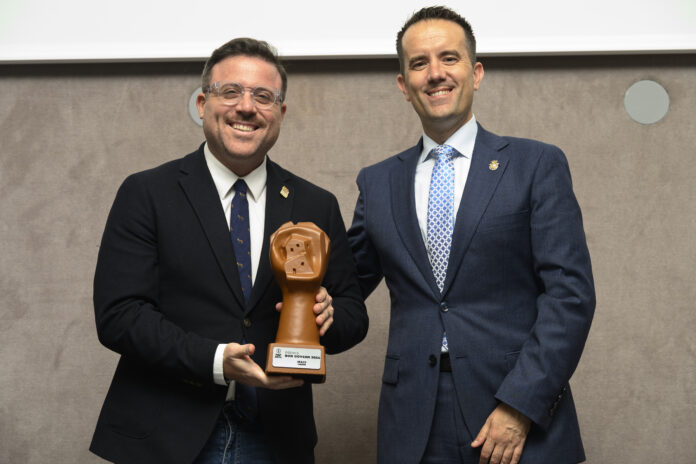 Two men in business suits smiling while holding a large bronze hand-shaped trophy.