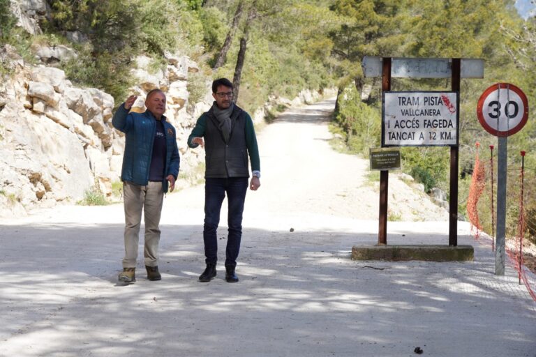 Two men stand on a dirt road beside a trail sign and a 30 km/h speed sign, with rocky hillside and trees in the background.
