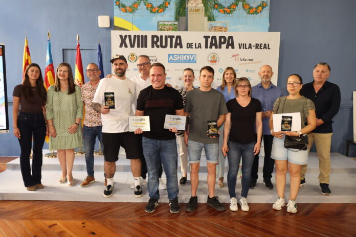 Group of people posing on stage at a festival event with a banner reading XVIII Ruta de la Tapa Vila-real in the background on a blue wall behind them, some holding flyers.