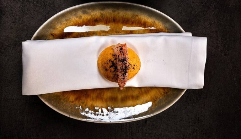 Overhead view of a plated appetizer: a round orange croquette topped with a strip of crispy bacon on a white napkin, placed on a glazed ceramic plate.