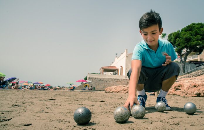 Boy in a turquoise polo crouches on the sandy beach placing metal boules for a game, with colorful umbrellas in the background.