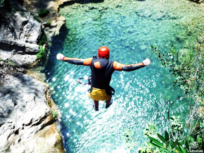 Person in an orange wetsuit and red helmet jumps from a rocky ledge into bright turquoise water.
