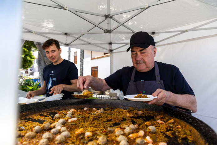 Two men cook paella outdoors under a white canopy, scooping rice and seafood from a large pan.