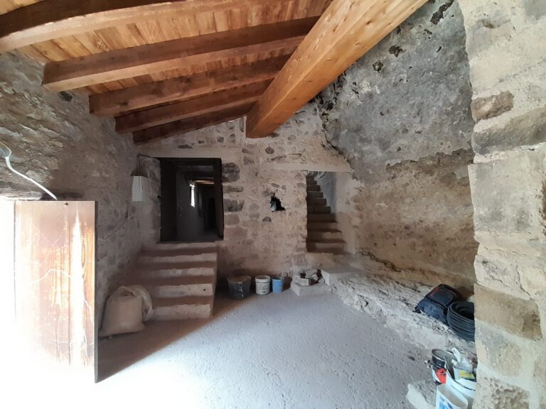 Stone basement with rough stone walls, exposed wooden ceiling beams, and a stairway descending to a lower level; buckets and construction gear on the floor.