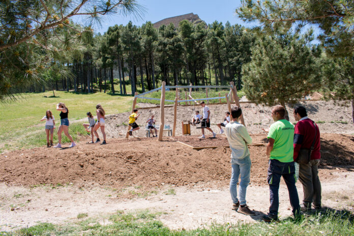 Children play on a wooden playground frame as adults supervise in a sunny park with pine trees and a hill in the background.