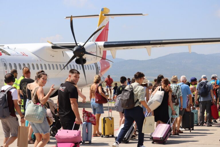Crowd of travelers with colorful suitcases lined up on a tarmac beside a propeller airplane, awaiting boarding.
