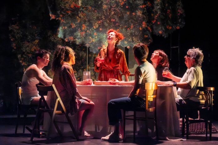 A woman in orange robe stands and sings at a round table surrounded by seated cast members holding drinks, in a dim stage setting with a leafy backdrop.