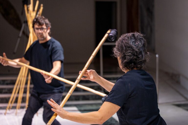 Two people practicing with long bamboo poles in a dim, spacious studio or rehearsal area.