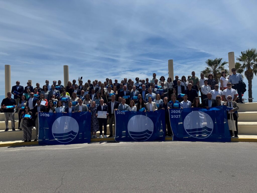 Large group of people posing on steps outdoors, holding a blue banner with 2026/2027, under a clear blue sky with palm trees in the background.