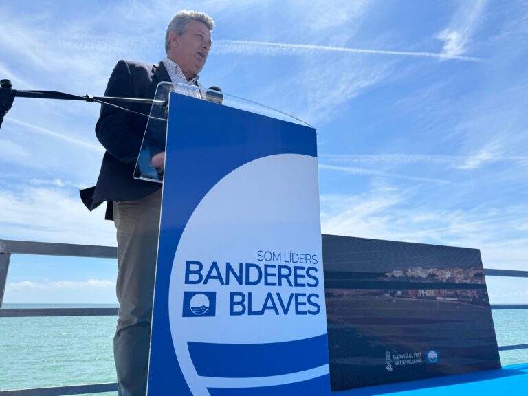 Man giving a speech at an outdoor podium by the sea, with blue and white banners and logos in the foreground.