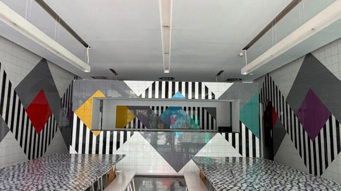 Modern cafeteria or dining area with a bold geometric mural: black-and-white vertical stripes, colored diamonds, and gray triangles; two long patterned tabletops with white benches.