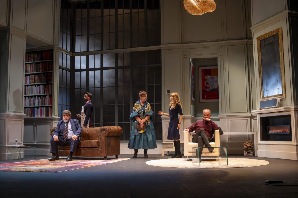 Actors on a theater stage in a living-room set: tall windows, bookshelves, sofa, chairs, fireplace, and modern decor.